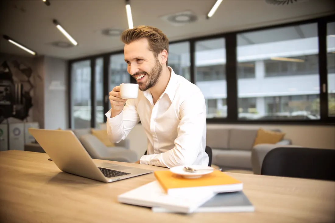 Mann im hellen Büro trinkt lächelnd einen Kaffee am Laptop